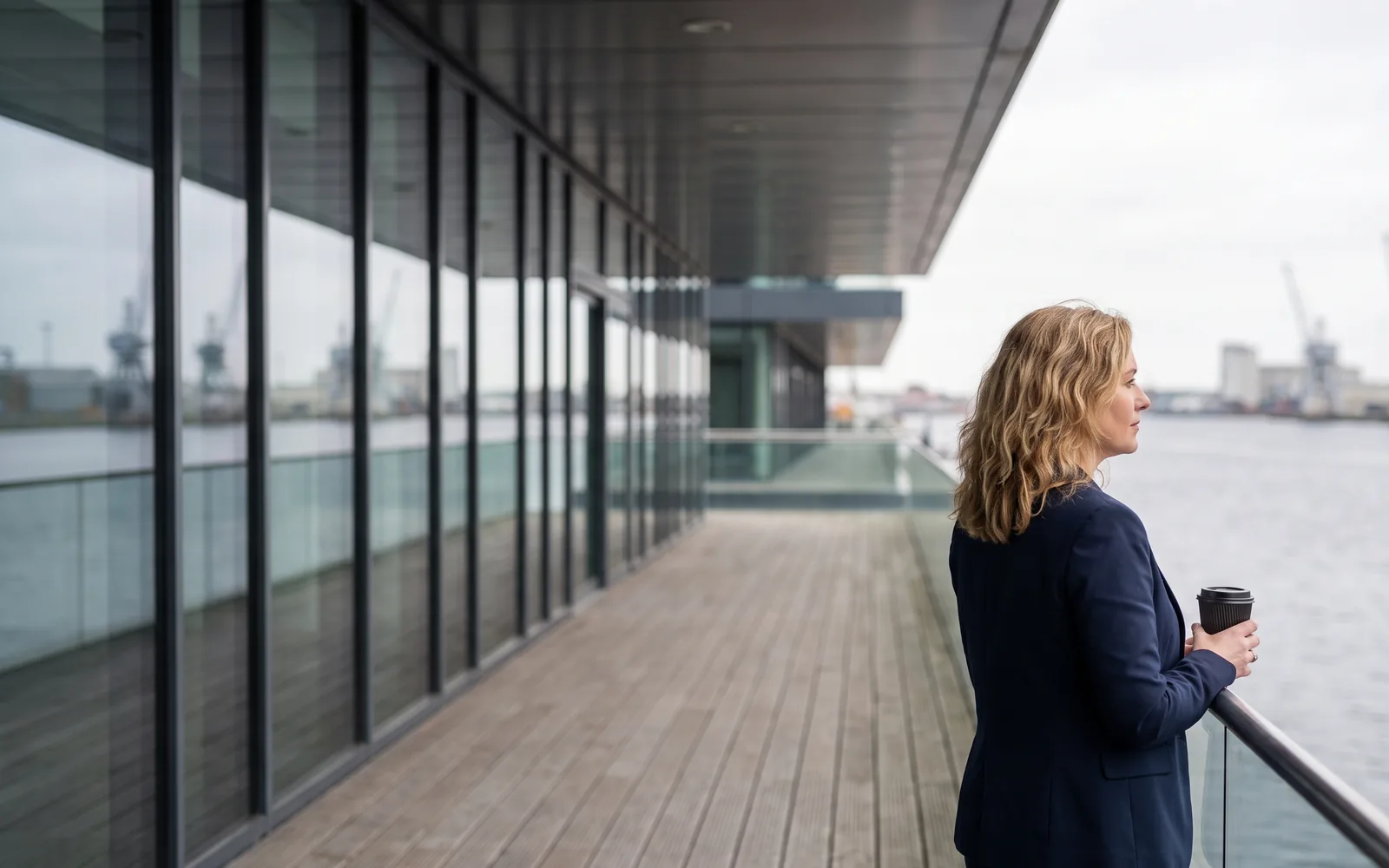 An executive in a navy blazer holds a coffee on a covered dockside office terrace, looking out over grey water